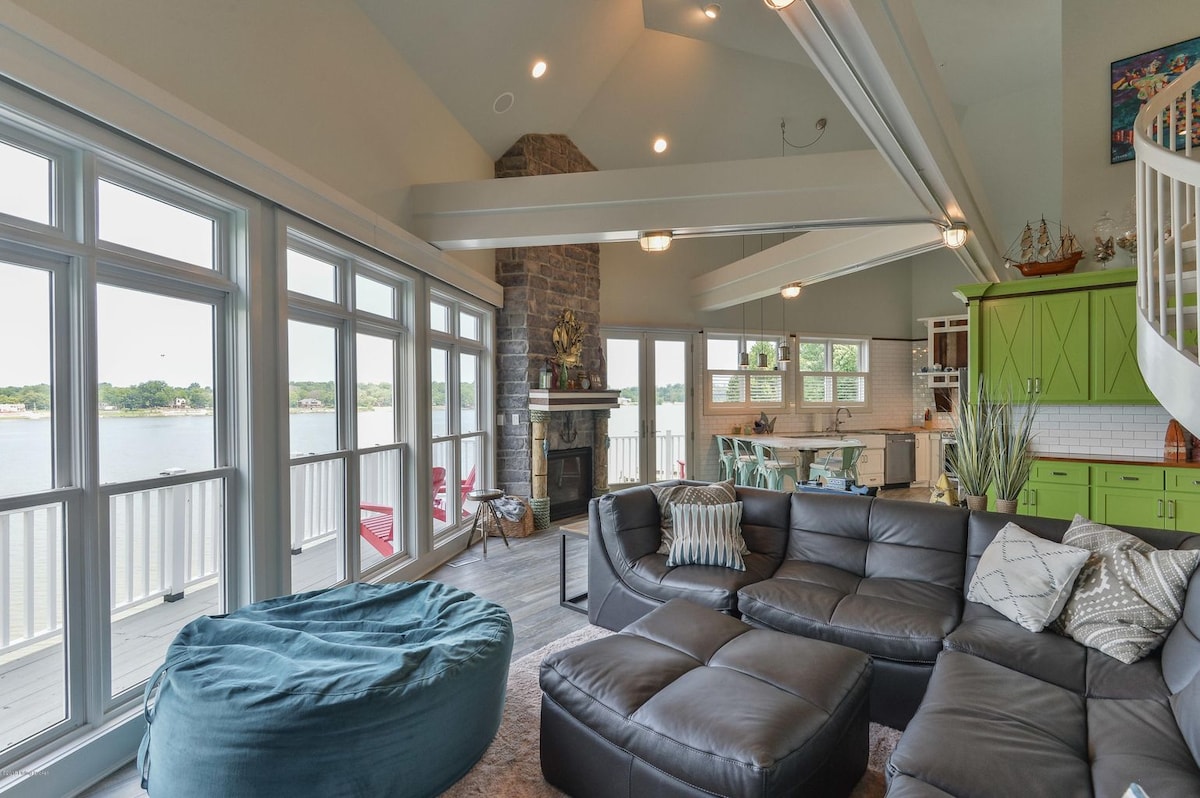 A spacious living area is highlighted by large windows offering panoramic views of the waterfront. A modern sectional sofa and a beanbag chair provide ample seating, while natural light floods the room. The decor features a mix of green cabinetry and stone accents, creating a casual yet stylish atmosphere.