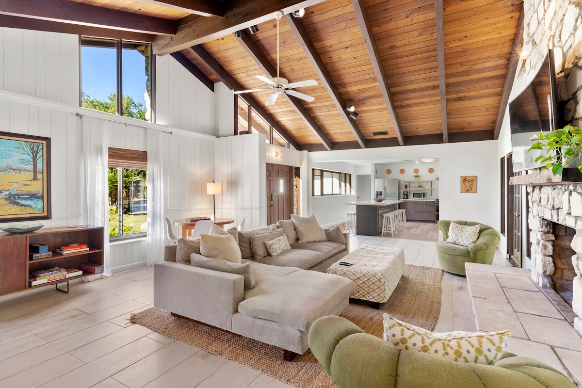 A spacious living area features vaulted wooden ceilings and large windows that allow natural light to enter. A sectional sofa and accent chairs are arranged invitingly around a coffee table. A stone fireplace and wooden shelving add character to the room, along with art and plants.