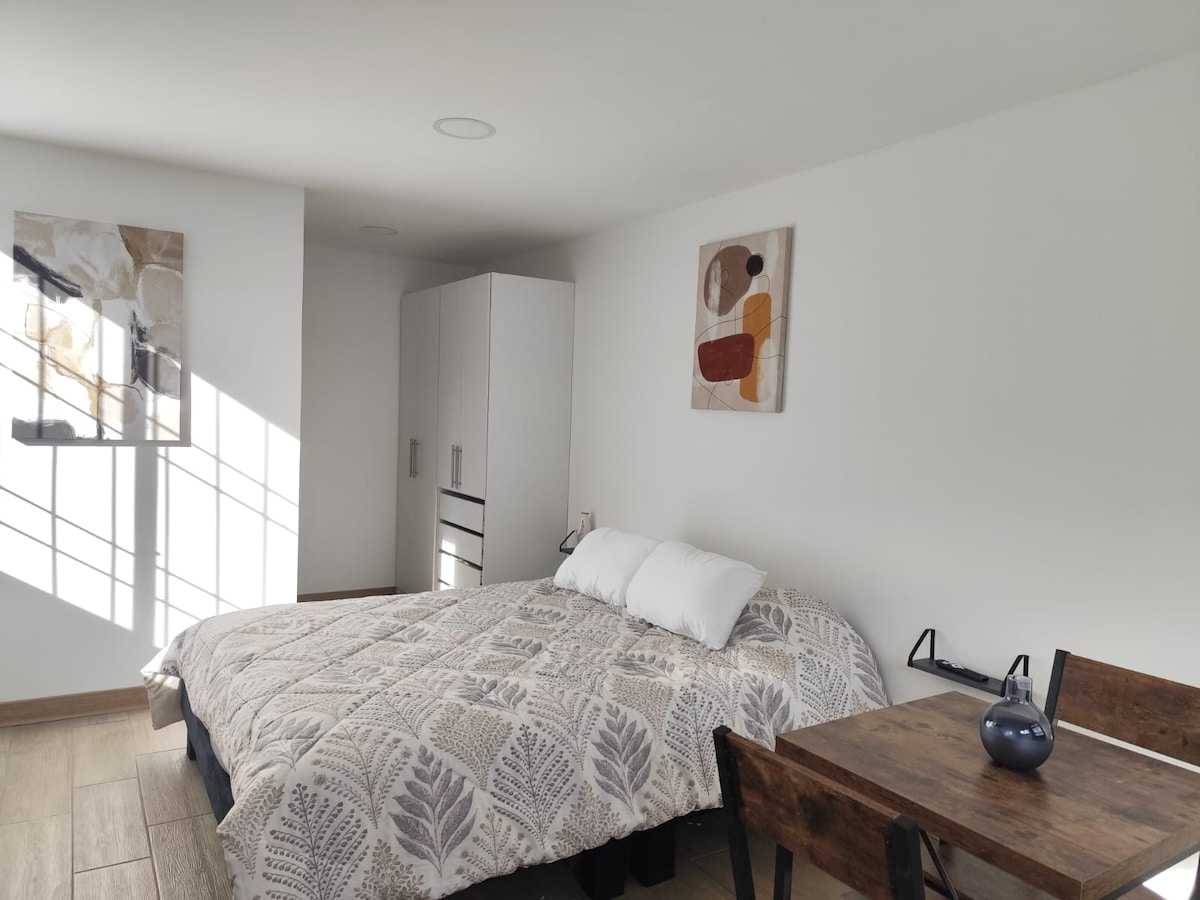 A spacious studio features a neatly made bed adorned with a patterned quilt and two decorative pillows. A streamlined cabinet stands nearby, complemented by a wooden table set for two. Soft light filters through a nearby window, creating a calm environment.