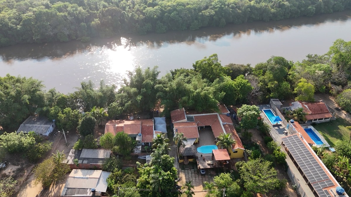 An aerial view captures the expansive layout of the property along the riverbank, surrounded by lush greenery. Several buildings are visible, along with two swimming pools reflecting the sunlight. The tranquil waters of the river complement the peaceful natural setting.