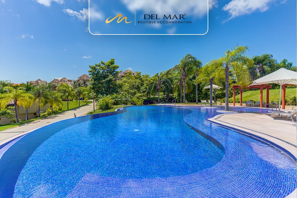 A striking infinity pool is framed by lush greenery and palm trees, offering a serene atmosphere. Sun loungers are arranged around the pool, inviting relaxation. The clear blue water reflects the bright sky above, enhancing the tranquil environment.