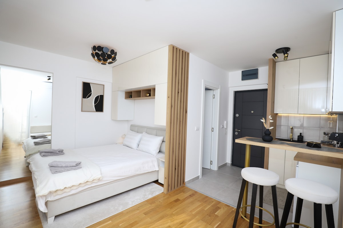 A well-furnished studio apartment features a spacious bed fitted with soft linens and pillows. The minimalist kitchen bar is equipped with modern stools, while light wood accents add warmth to the space. A doorway leads to a light-filled area in the background.