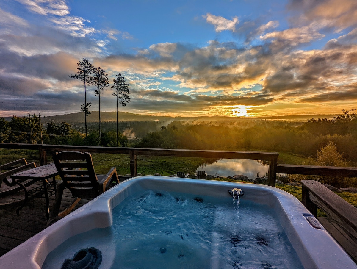 A hot tub is positioned on a wooden deck, overlooking expansive views of rolling hills and a tranquil pond. The sky displays a blend of soft colors during sunset, creating a serene atmosphere. Nearby, outdoor chairs are visible, inviting relaxation.