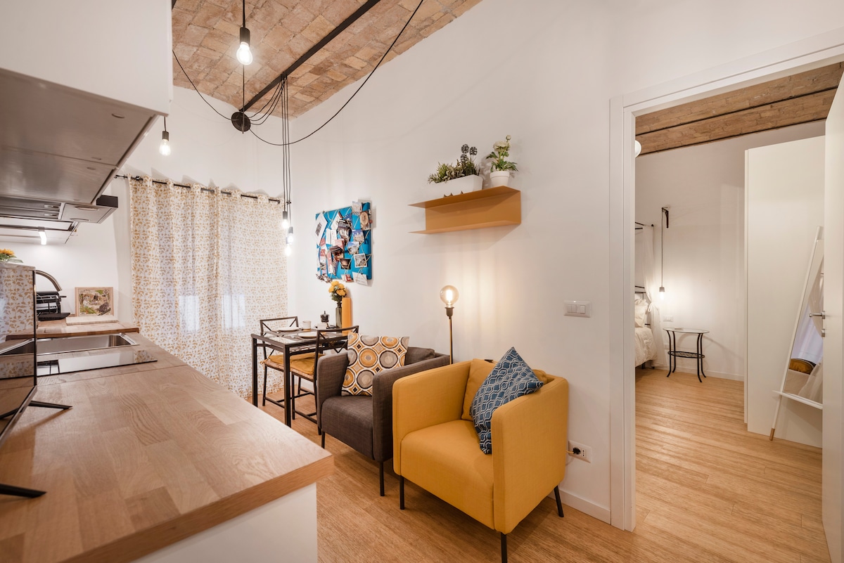 The image shows a bright open space featuring a dining nook with a wooden table and four chairs. Two cozy armchairs in yellow and gray are positioned nearby. A decorative wall shelf with a plant adds charm, while natural light filters through patterned curtains.