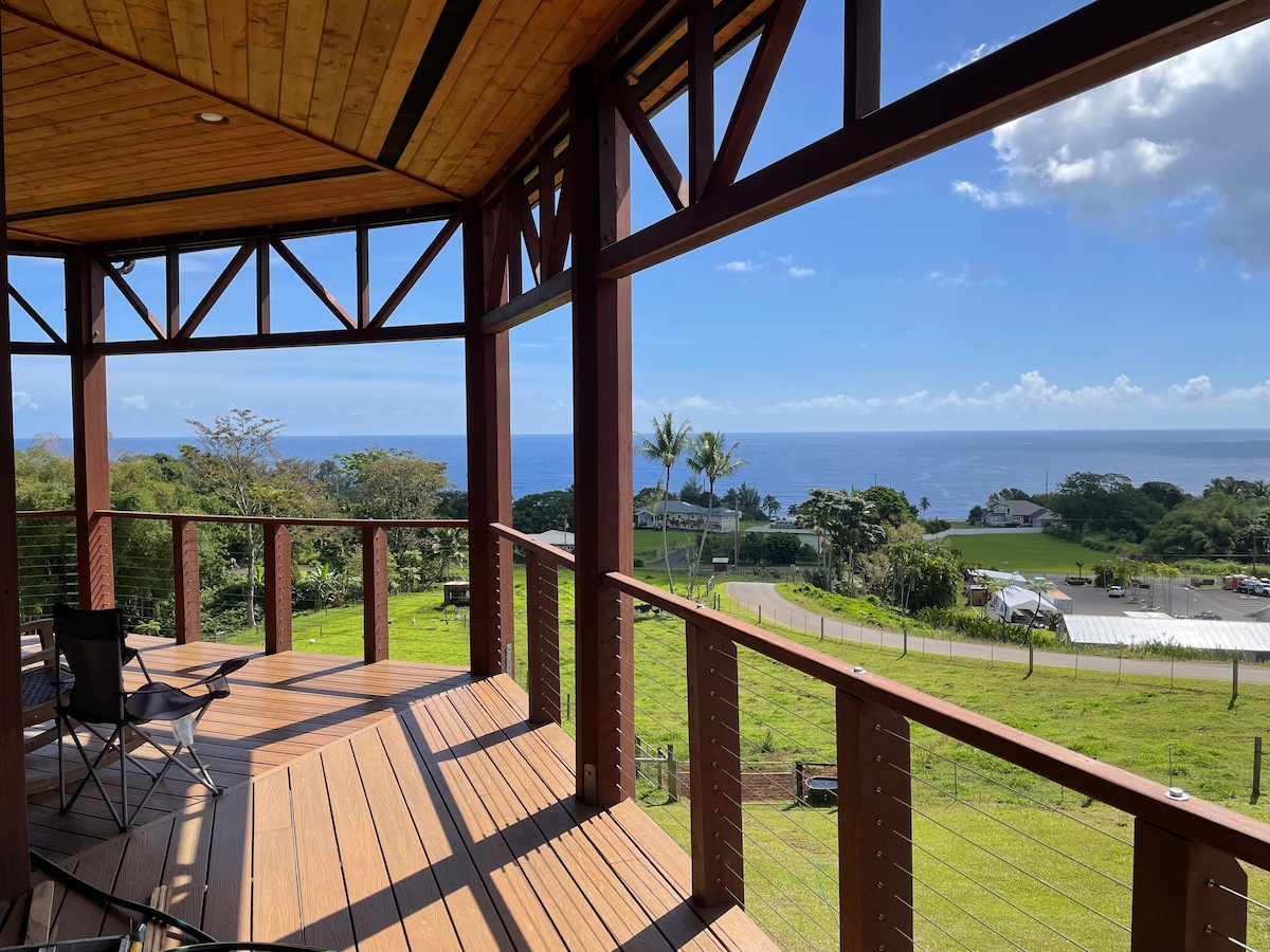 The spacious lanai offers unobstructed views of the ocean and surrounding greenery. The wooden structure features a blend of beams and posts, creating an open and airy feel. Two folding chairs are positioned to allow guests to enjoy the serene outdoor environment.