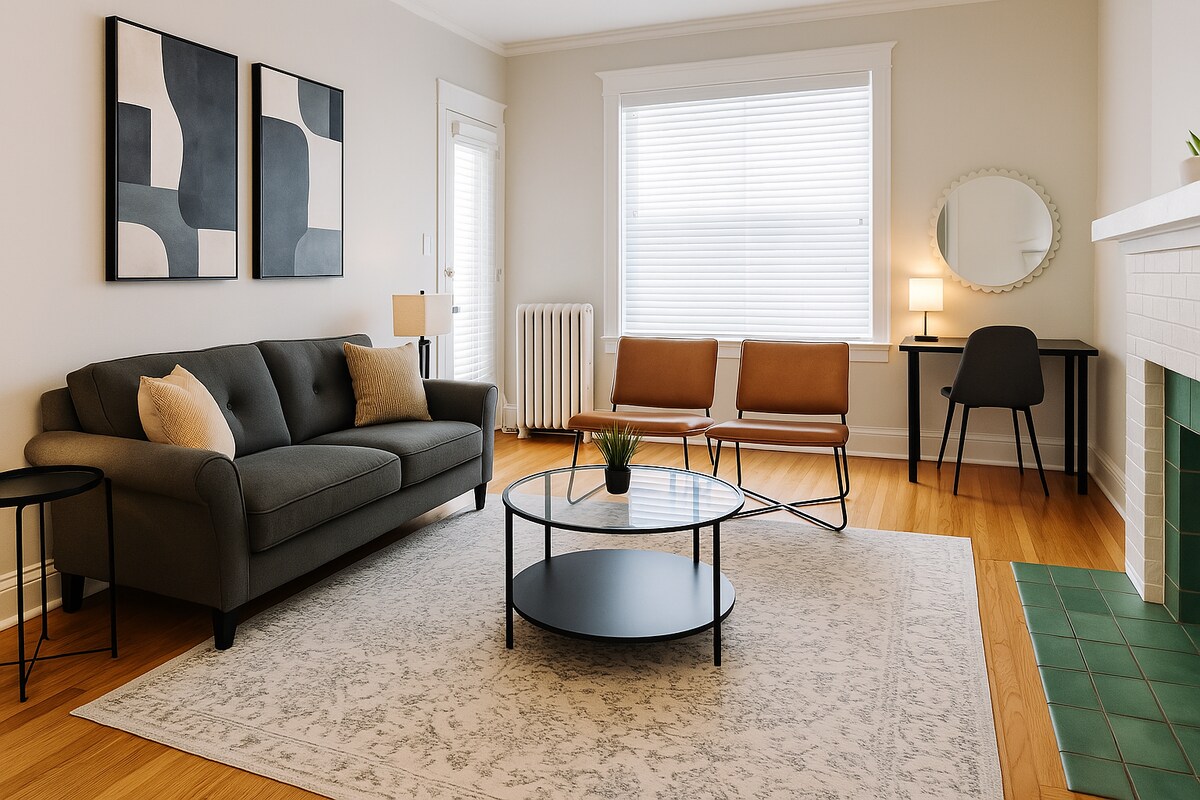 A modern living area features a dark gray sofa with accent pillows, a round coffee table, and mid-century style chairs. A decorative area rug lies beneath, while natural light enters through a large window with white blinds. A small desk and mirror are positioned against the wall.