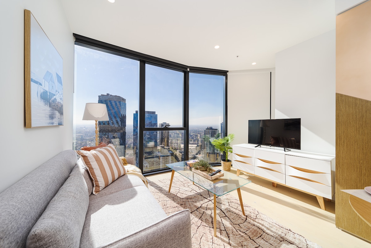 A modern living area is presented with large windows offering a panoramic view of the Melbourne skyline. A sleek white cabinet holds a television, while a glass coffee table is placed atop a patterned rug. A light grey sofa with accent pillows enhances the space.