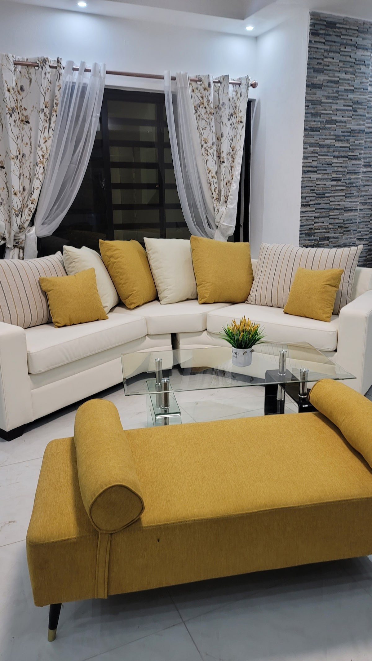A modern lounge area features a white sectional sofa adorned with assorted cushions in soft tones. A glass-top coffee table sits in front, complemented by a small decorative plant. Natural light filters through sheer curtains, illuminating the sleek stone wall behind.