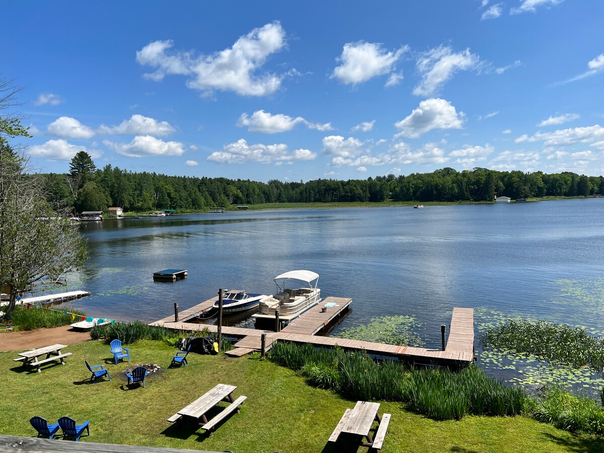 A scenic view of Catfish Lake is captured, featuring a wooden dock with a boat and a paddle boat tied alongside. The lush green yard holds several picnic tables and colorful chairs, surrounded by gentle ripples of water reflecting a clear blue sky with fluffy white clouds.