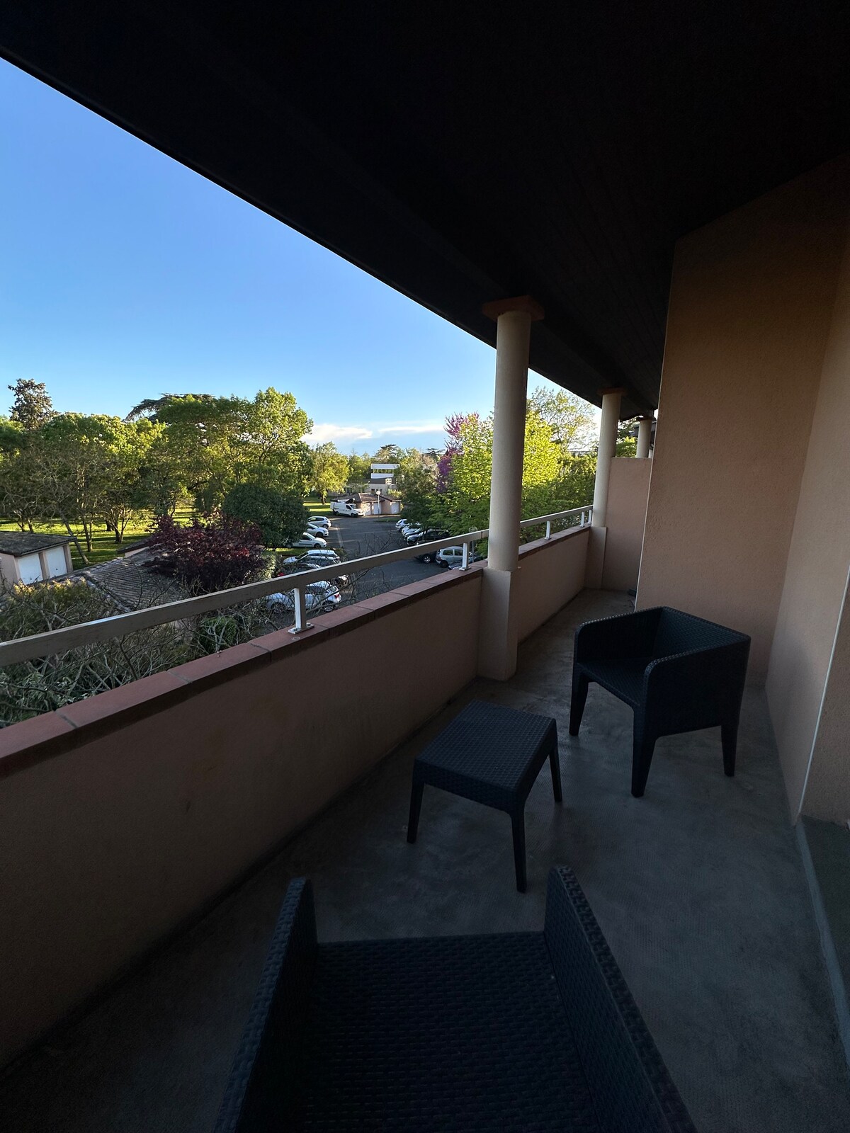 A private balcony features two black woven chairs with a view of the surrounding greenery and quiet streets. The bright sky indicates pleasant weather, and the structure's walls provide a sense of enclosure while allowing for outdoor relaxation.