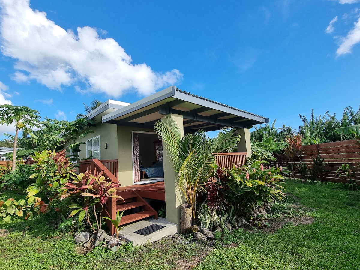 A tropical bungalow is set amidst lush greenery, featuring a covered porch with wooden steps leading to a vibrant garden. Bright blue skies and scattered clouds provide a serene backdrop. The structure is complemented by a natural landscape with various plants and foliage surrounding it.