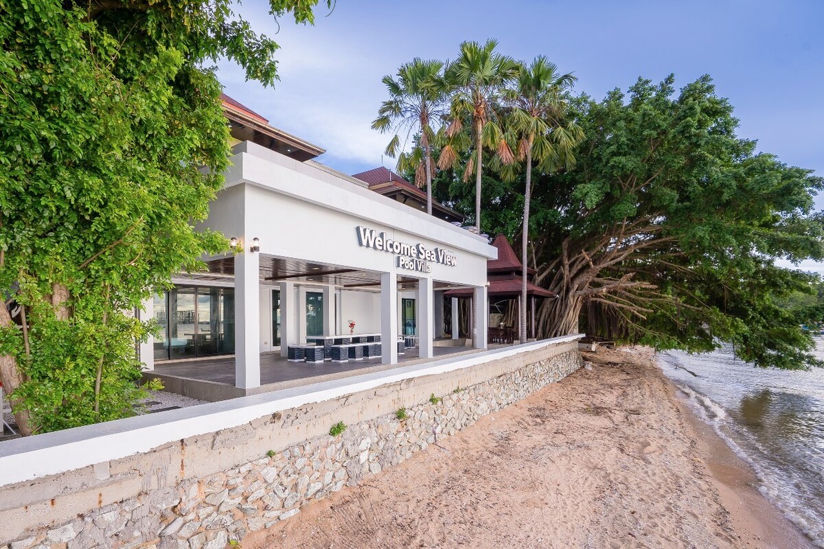 A beachfront villa is showcased, featuring a modern architectural design with large windows. Lush palm trees frame the property, and sandy beach leads to the water. The welcoming sign is prominently displayed above the entrance, indicating a serene environment.