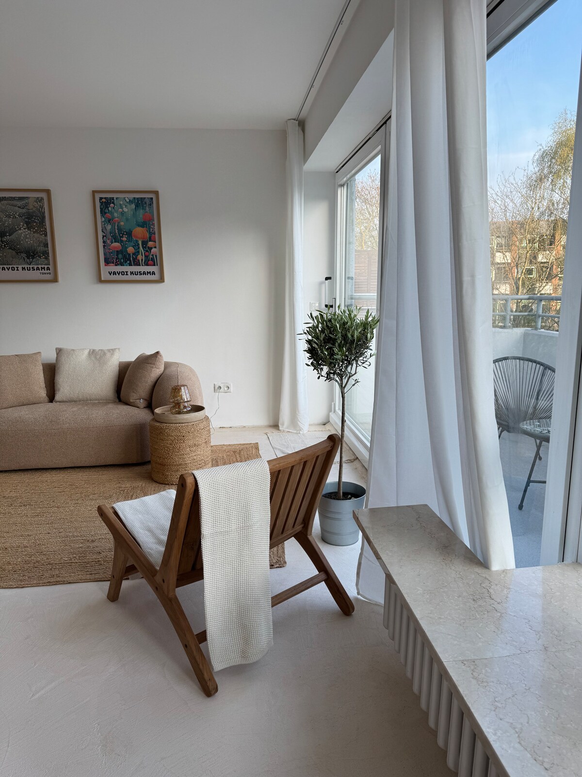 A well-lit living area features a cozy beige sofa and a wooden folding chair accented with a white throw. Light curtains frame large glass doors leading to a balcony, allowing natural light to fill the space. Decorative artwork is displayed on the wall.