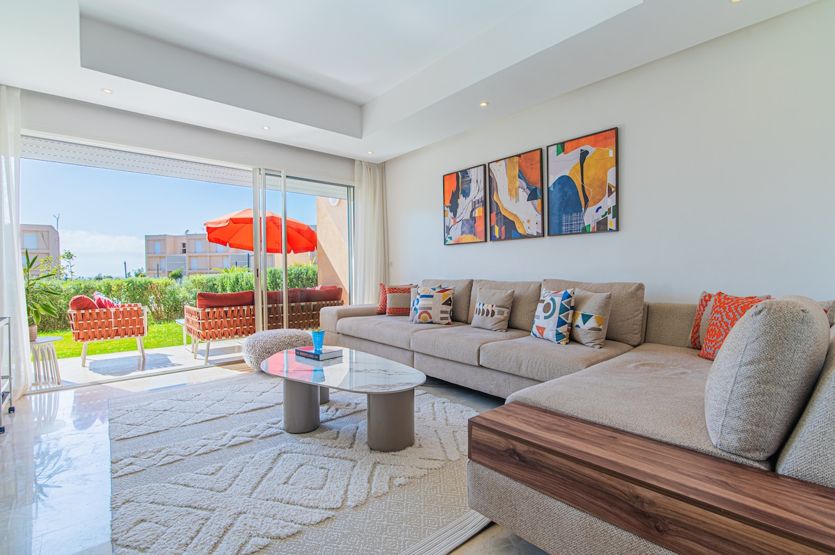 A spacious living area features a comfortable sectional sofa adorned with colorful cushions. Large glass doors open to a garden view, allowing natural light to brighten the room. A modern coffee table sits on a textured rug, complemented by vibrant artwork on the walls.