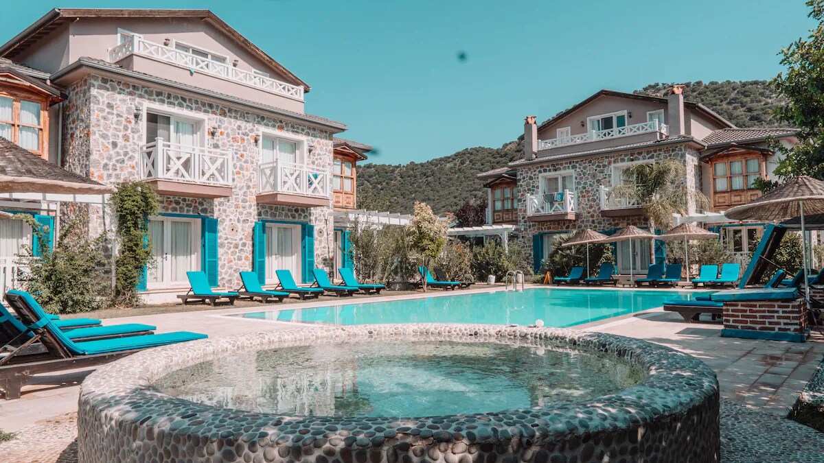 A refreshing swimming pool is surrounded by lounge chairs in a tranquil outdoor area. The building features a natural stone facade and balconies. Umbrellas provide shade for guests, and the lush hills in the background add greenery to the scenic view.