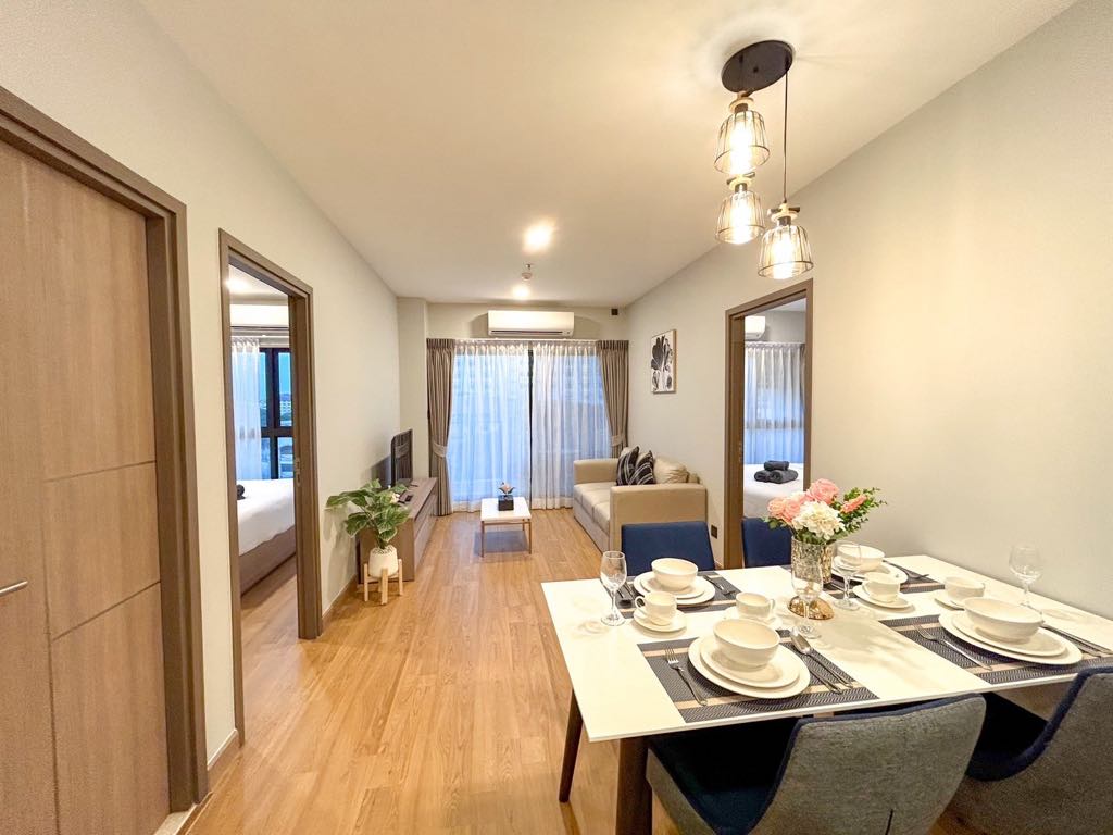 A modern dining area is showcased, featuring a table set for four, with elegant plates and cutlery arranged neatly. A cozy living space is visible, adorned with a sofa and a smart TV. Natural light filters through the large windows, enhancing the inviting ambience.