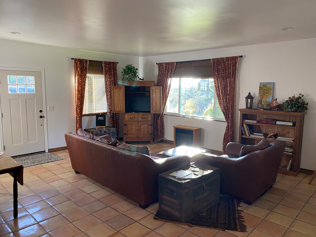 A living area is presented with a spacious leather sofa and a wooden coffee table. Two large windows are dressed with patterned curtains, allowing natural light to illuminate the tiled floor. A wooden cabinet holds a television, complemented by decorative elements on shelves.