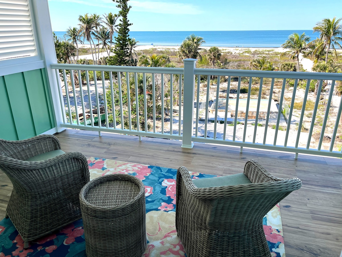 A serene balcony view features two woven chairs facing the ocean, with a small table between them. The floor is adorned with a colorful rug, while the backdrop showcases palm trees and a beach extending towards the horizon under a clear blue sky.