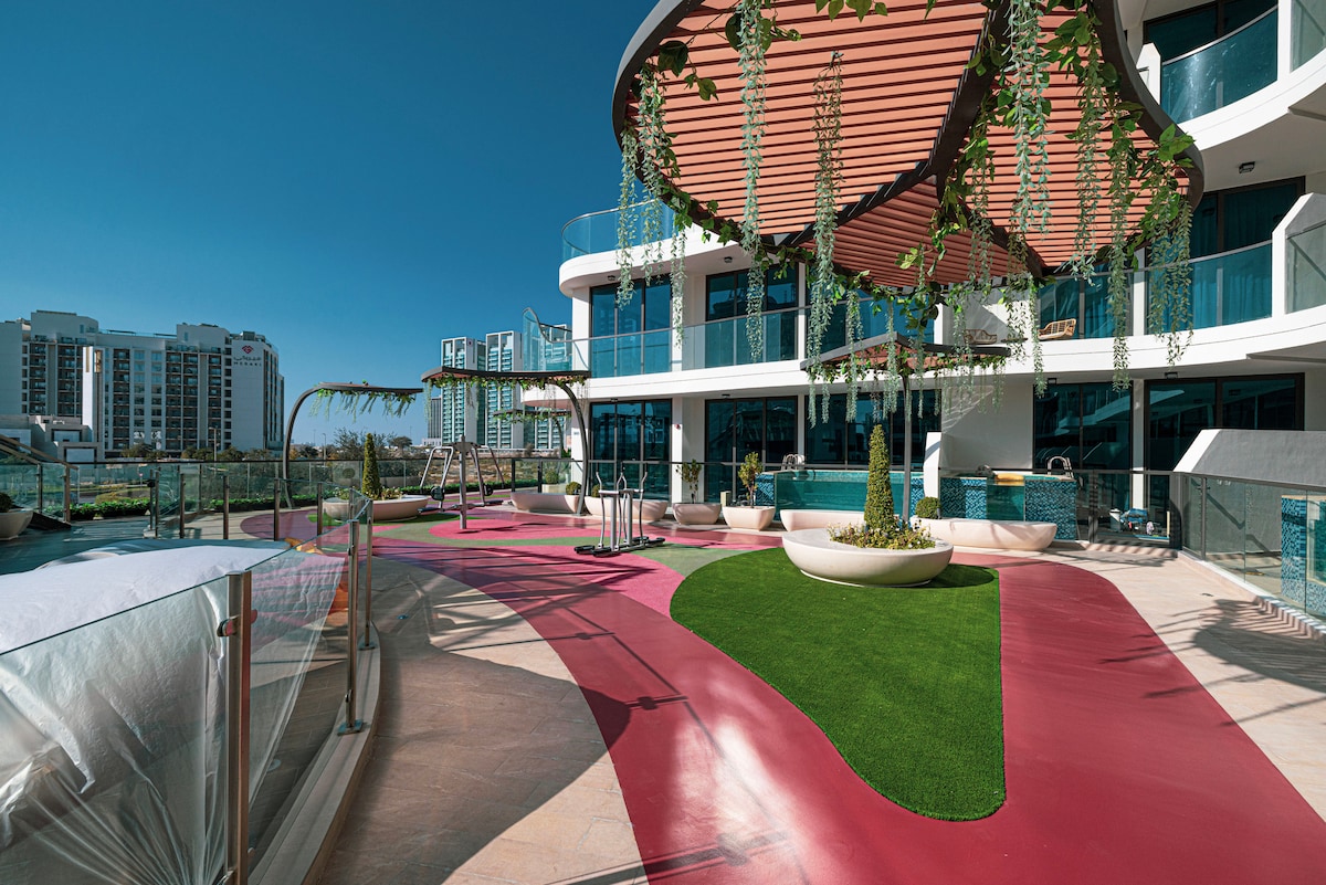 A spacious outdoor terrace features modern seating areas, lush greenery, and a vibrant pathway. Sunlight reflects off the glass structure in the background, enhancing the inviting atmosphere. The area includes a swimming pool and multiple lounging spaces for relaxation.