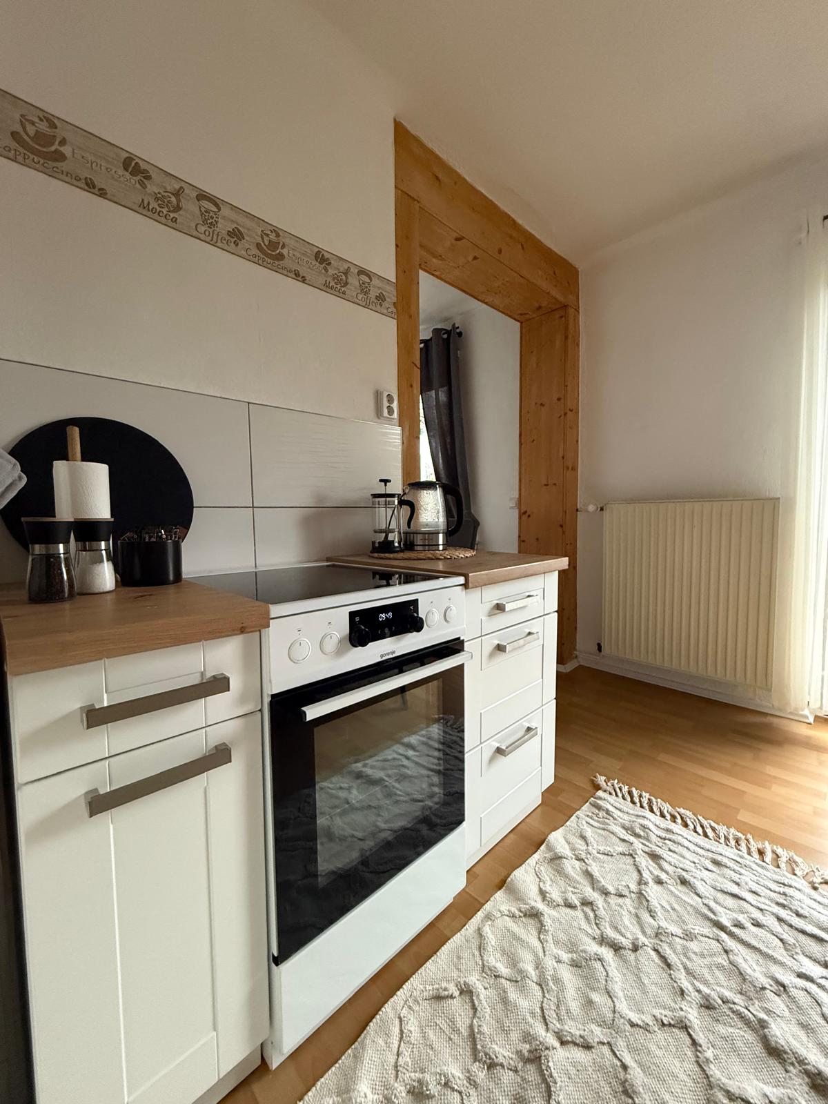The compact kitchen is equipped with modern appliances, including an oven and a stovetop. Light wood cabinetry contrasts with the white tiles and countertop. A cozy rug lies on the floor, and a window allows natural light to illuminate the space.