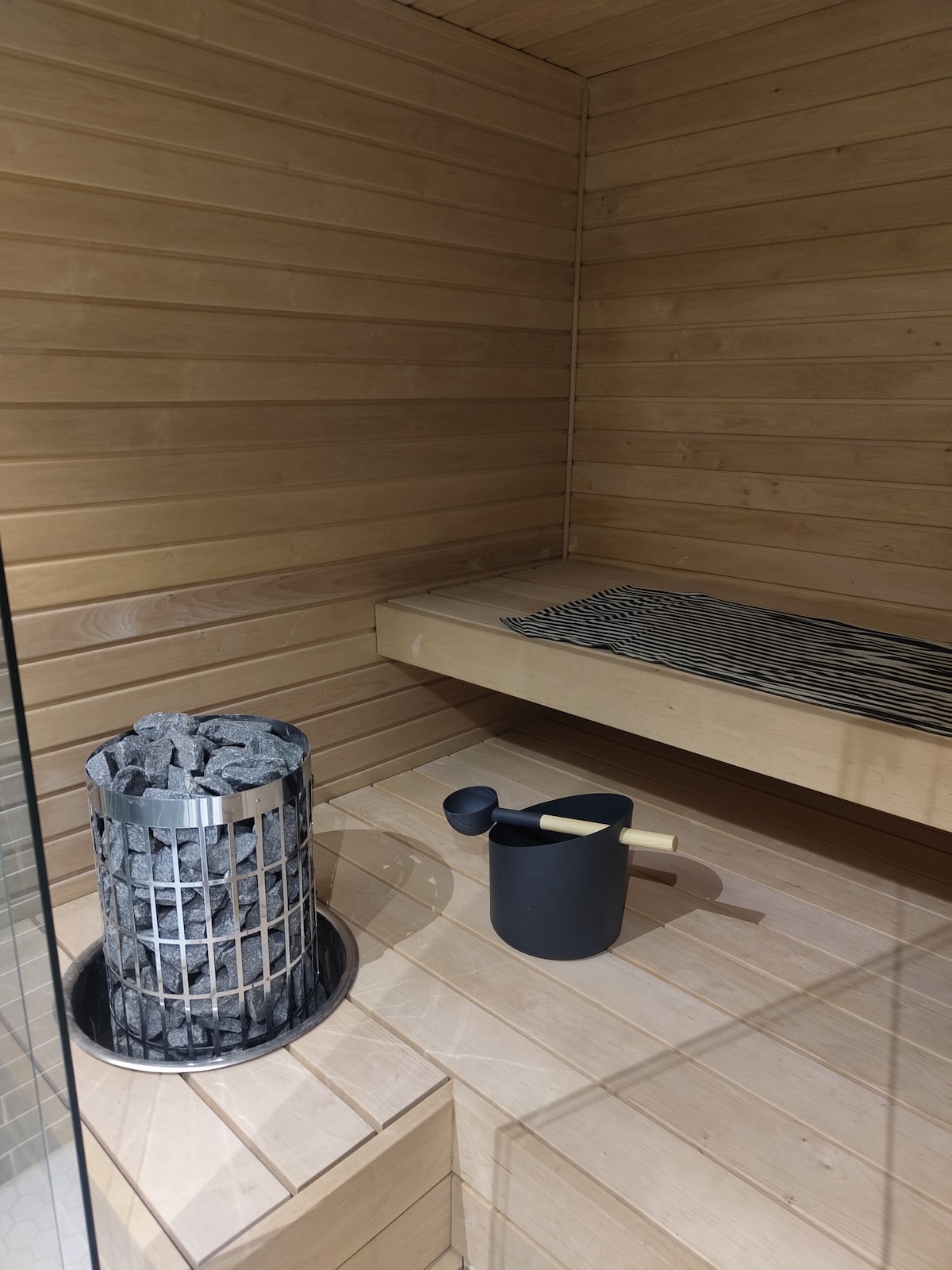 The image captures a serene sauna environment characterized by warm wooden walls and benches. A stone heater stands prominently, surrounded by a metal basket filled with rocks. A small black bucket and ladle sit nearby, complementing the relaxing atmosphere.