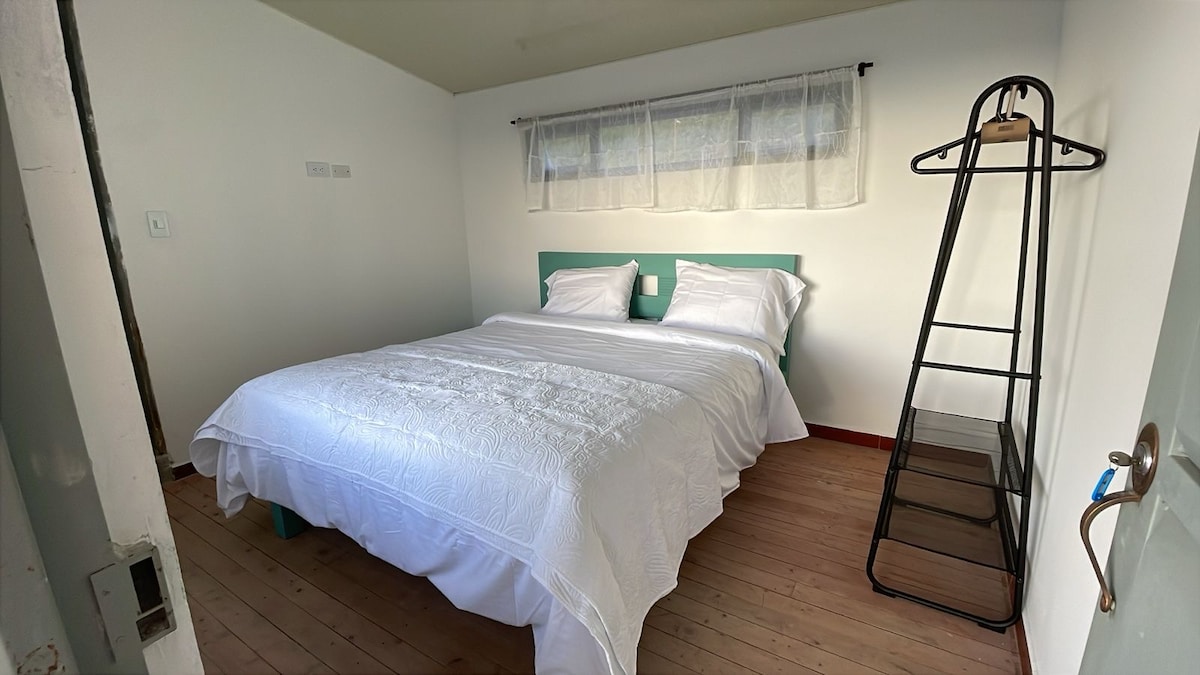 A neatly arranged double room is featured, promoting a calming atmosphere with a spacious bed topped with white linens. A minimalistic clothing rack stands beside the bed, complementing the light-colored walls and wooden flooring. Natural light filters through a sheer curtain at the window.