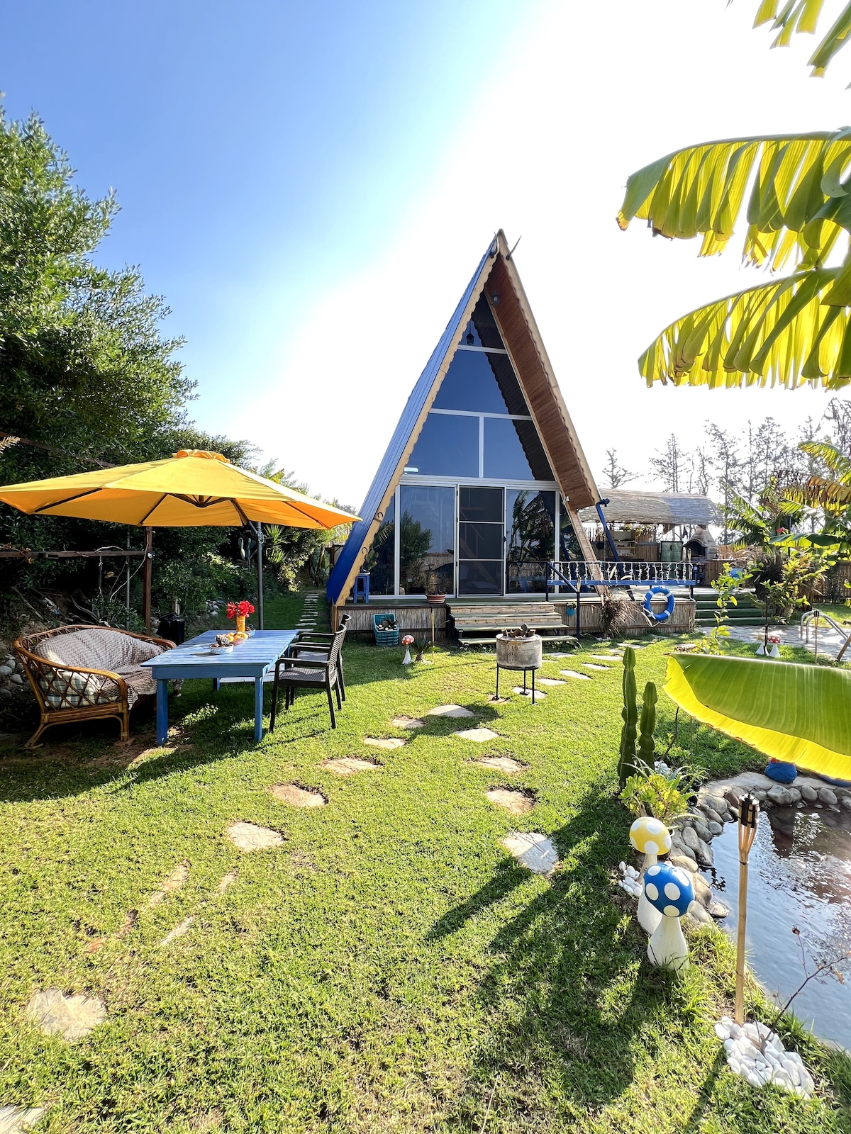 An A-frame house is prominently situated amidst greenery, featuring large glass windows that invite ample natural light. A yellow umbrella shades a dining area with a wooden table and chairs. A pathway of stones leads to a tranquil pond adorned with decorative elements.