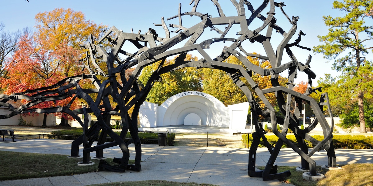 An artistic archway crafted from intertwined black branches frames a white circular stage. Surrounding landscape features fall foliage with vibrant colors in the background, contributing to a serene outdoor setting.