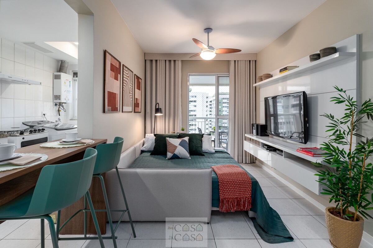 A combined living and dining area is presented, featuring a cozy futon with decorative pillows. A small dining table with high-backed chairs is visible, along with a TV mounted on a white entertainment unit. Natural light streams through a window, complemented by soft beige walls.