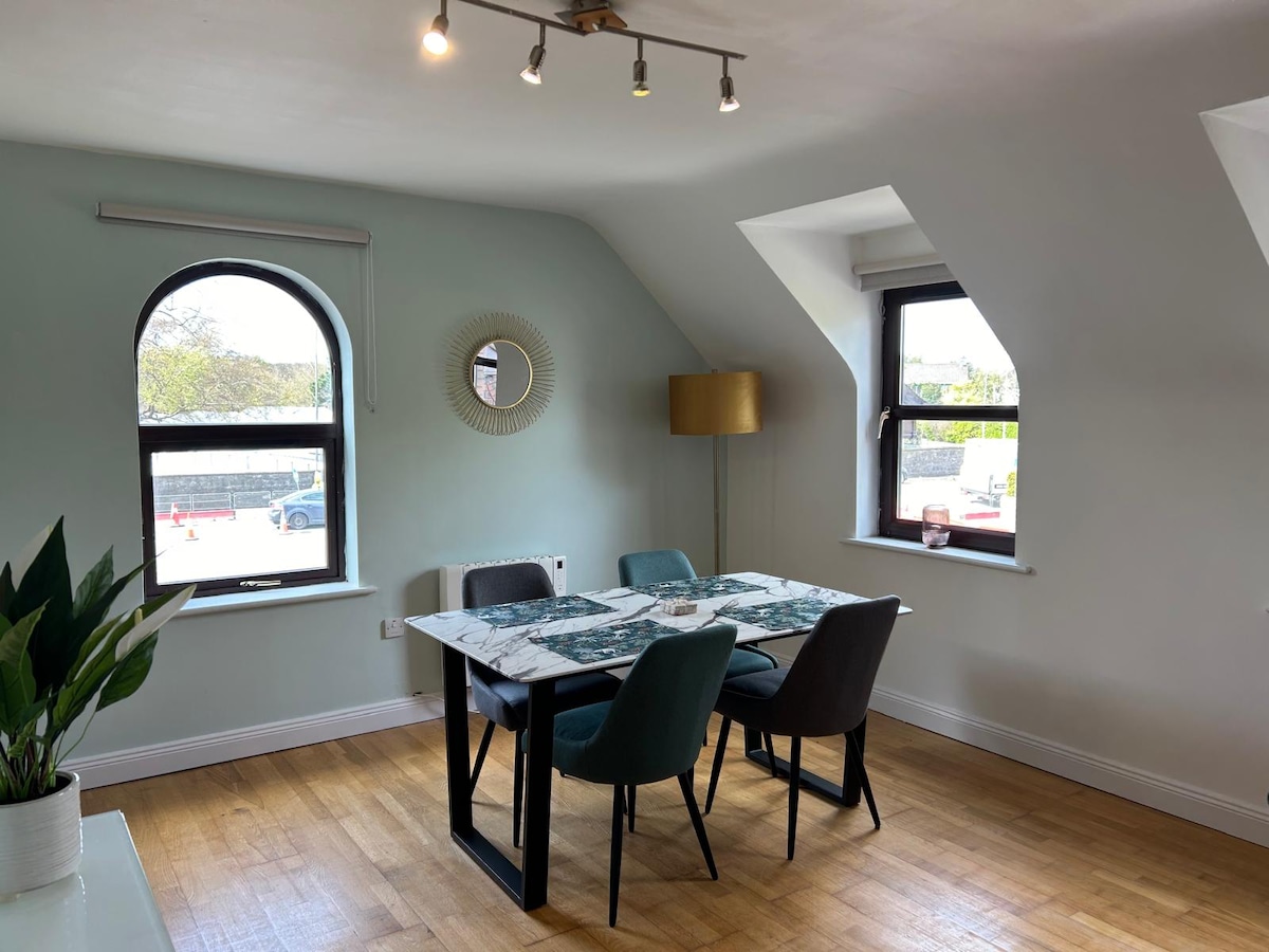 A dining area is presented with a rectangular table surrounded by four chairs. Natural light enters through two large windows, highlighting the simple decor. A round mirror is mounted on the wall, and a lamp stands nearby, contributing to a bright and functional space.