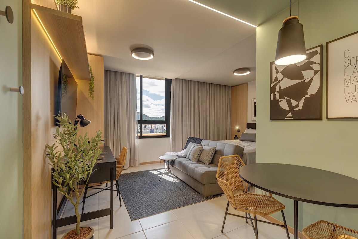 A modern living space features a cozy seating area with a gray sofa and decorative plant. A round dining table is surrounded by two woven chairs. Light-colored walls and large windows offer natural light, while abstract wall art adds a stylish touch to the room.