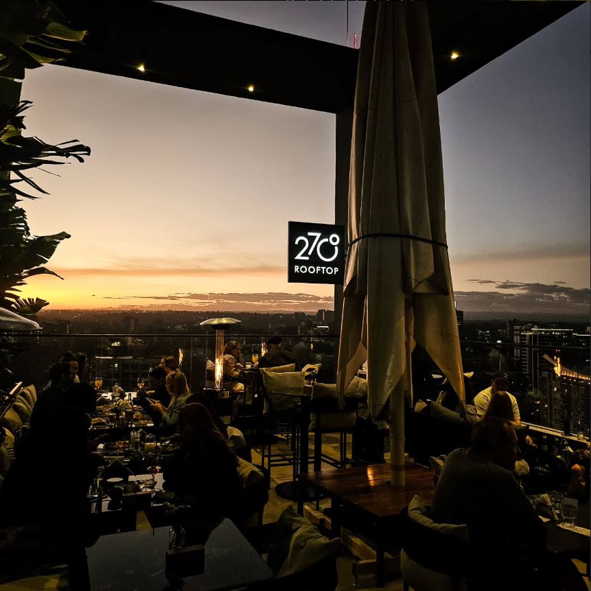 The rooftop dining area is illuminated by soft lighting as guests enjoy their meals, surrounded by tropical plants. A sign reading '278 Rooftop' is visible against the backdrop of a sunset, casting warm hues over the landscape and city skyline.