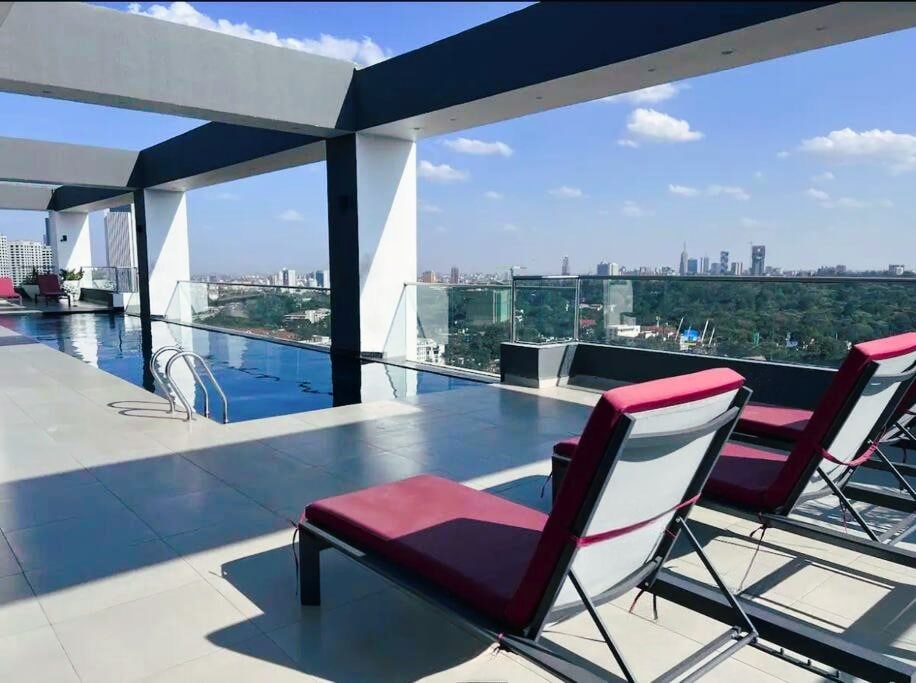 A sleek rooftop pool area offers stunning city views, accompanied by two lounge chairs positioned for relaxation. The expansive pool reflects the sky, with a backdrop of the skyline and open spaces that invite tranquility.