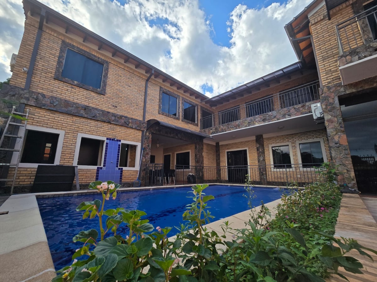An inviting outdoor space features a swimming pool surrounded by a deck. The building's stone exterior and balconies overlook the pool area, with several windows allowing natural light. Lush greenery borders the pool, enhancing the tranquil setting.