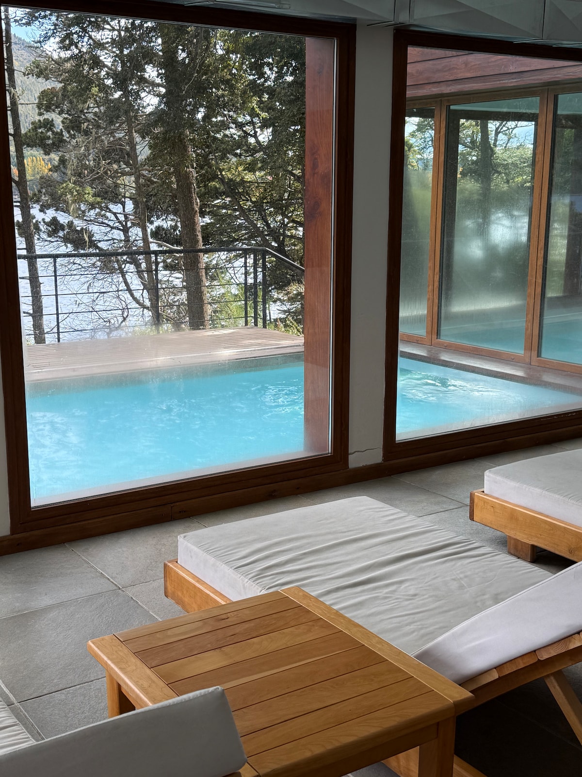 A serene indoor space features wooden loungers positioned near expansive glass windows, offering views of a tranquil pool and the surrounding nature. Natural light fills the area, highlighting the soothing blue water of the pool and the lush greenery outside.