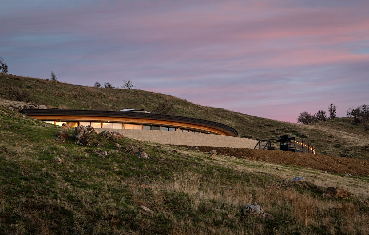 A modern architectural residence rests on a grassy hillside, framed by soft rolling terrain. Large windows reflect the evening light under a twilight sky, highlighting the seamless integration with the natural landscape. The gentle curve of the structure complements the serene surroundings.