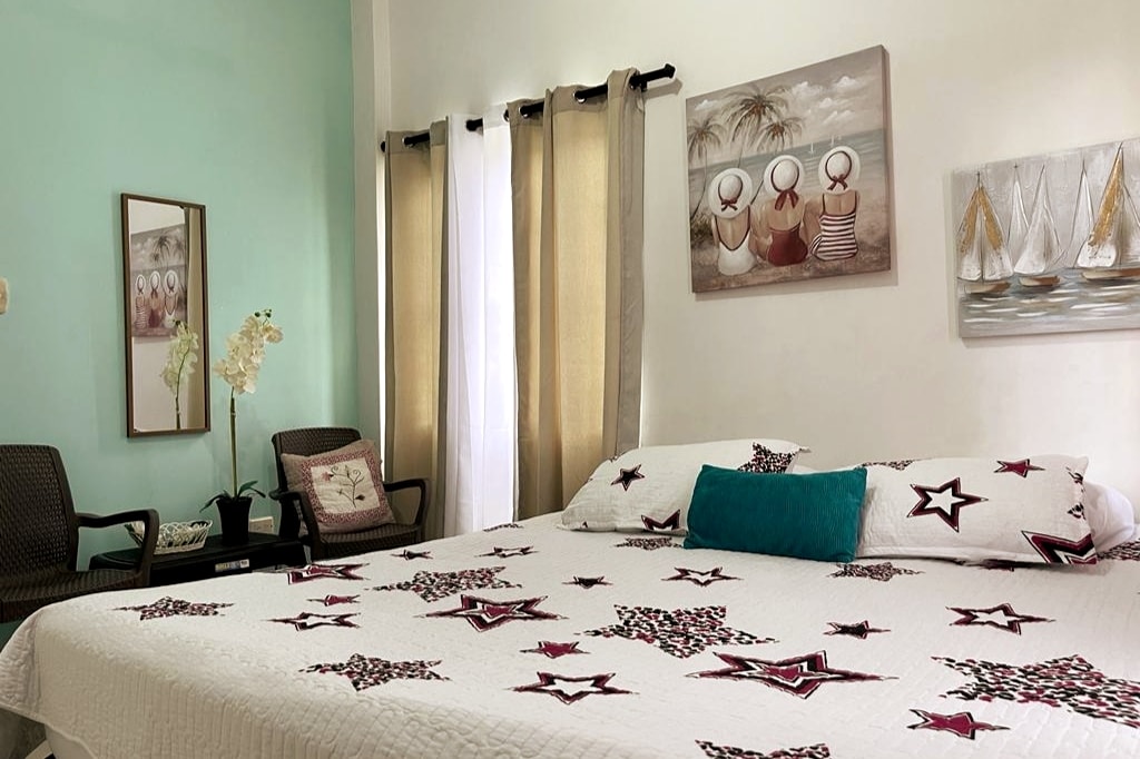 A cozy sleeping area is presented with a spacious bed featuring a star-patterned quilt. Two large windows are dressed with sheer white curtains and gold drapes, allowing natural light to fill the room. A chair and side table add a functional touch, complemented by wall art.
