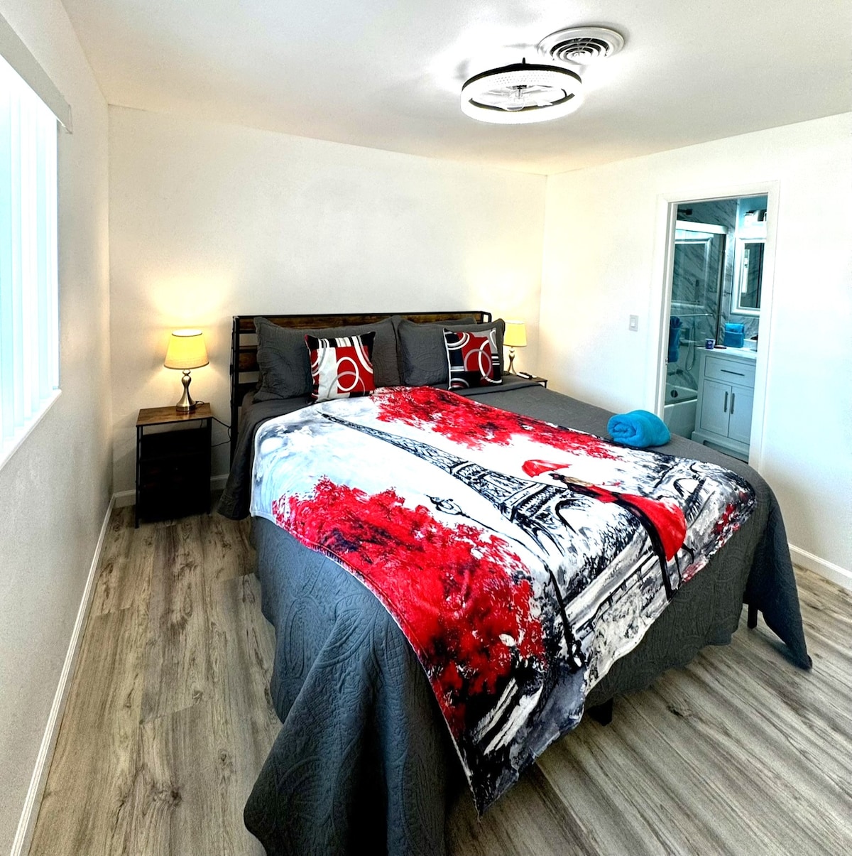 A cozy bedroom features a king-size bed adorned with a colorful red and black decorative blanket. Nightstands with lamps are positioned on either side, while a modern ceiling light illuminates the room. An attached bathroom is visible in the background.
