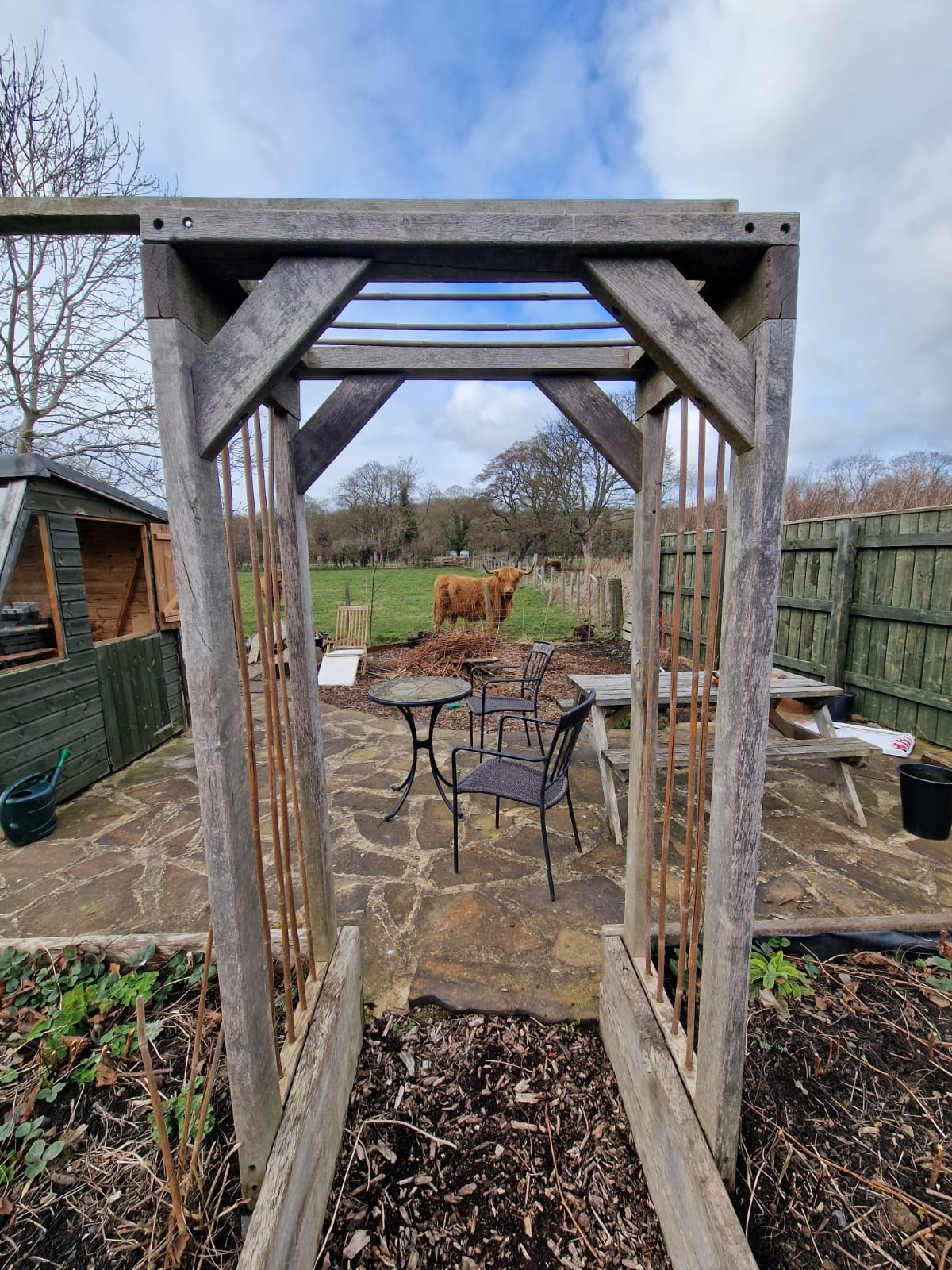 An archway constructed from wooden beams frames a view of a stone patio area with a small round table and two chairs. In the background, a highland cow is visible grazing, enhancing the outdoor garden's natural setting.