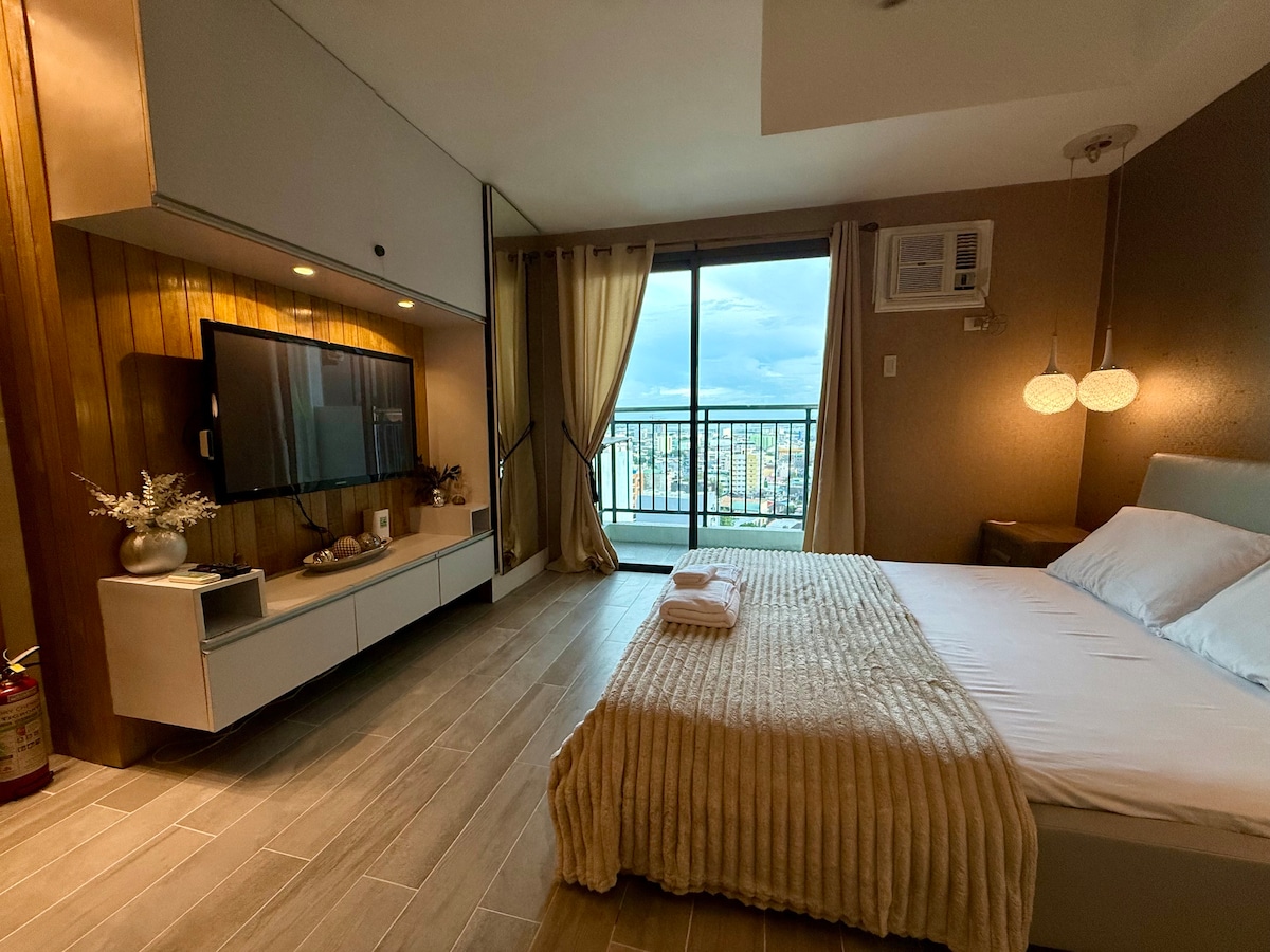 A modern bedroom featuring a spacious bed dressed in soft linens. A wall-mounted television is positioned across from the bed. Floor-to-ceiling windows provide views of the city, with sheer curtains allowing natural light to enter. An air conditioning unit hangs above the bed.