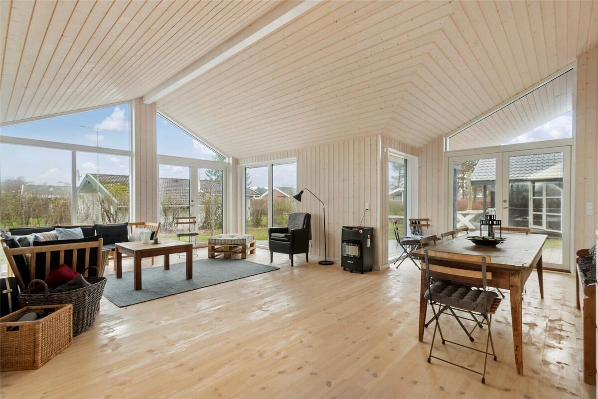 An open living space features high, sloped wooden ceilings and large glass windows, ensuring ample natural light. The area includes a dining table with several chairs, a comfortable seating arrangement, and a mixture of decorative elements. A warm wooden floor complements the overall design.