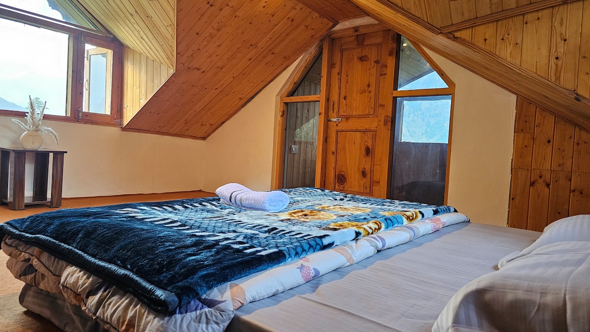 A cozy attic bedroom features a soft double bed adorned with a colorful blanket and a rolled towel. Wooden panels extend along the sloped ceiling, complementing the rustic charm. Large windows with wooden frames allow natural light to illuminate the room, offering views of the surrounding mountains.