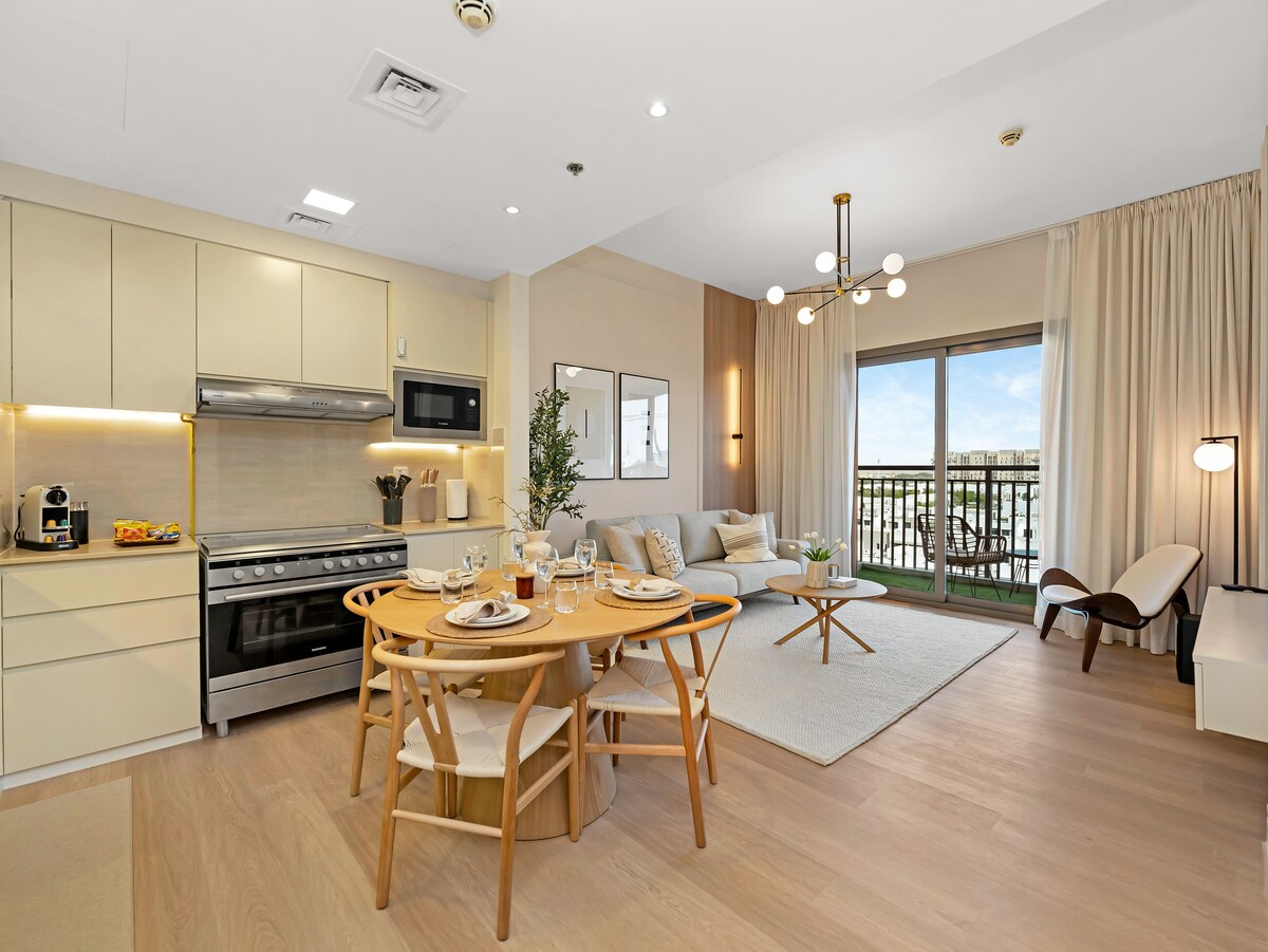 A bright and airy living-dining area is presented, featuring a round wooden dining table set for four. The modern kitchen is equipped with a stove and microwave. A comfortable sofa with soft cushions is positioned beside a large sliding door leading to a balcony with views.