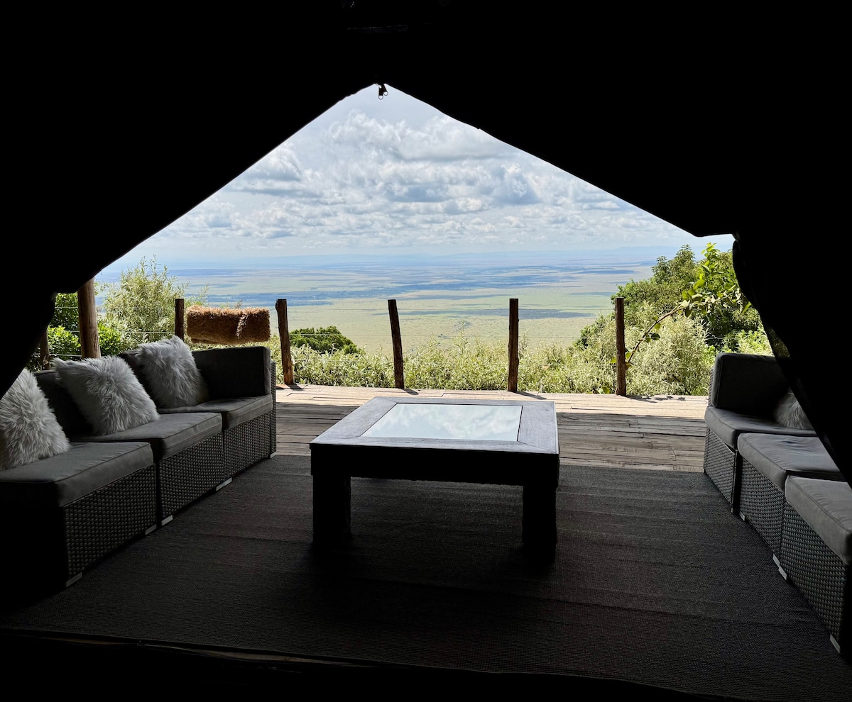 A spacious living area features comfortable seating arranged around a low coffee table. Large windows provide expansive views of the plains beyond, with natural light illuminating the interior. The flooring is covered with a textured mat, enhancing the cozy ambiance of the space.