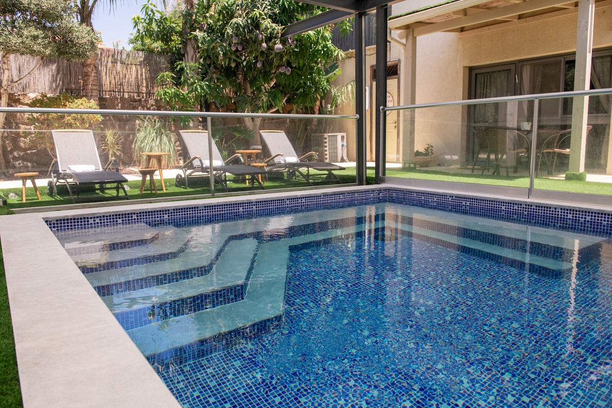 A private swimming pool showcases clear blue water surrounded by a modern patio. Comfortable lounge chairs are positioned nearby for relaxation. Lush greenery frames the area, enhancing the serene environment. The pool design includes varying depths and is accessible via steps.