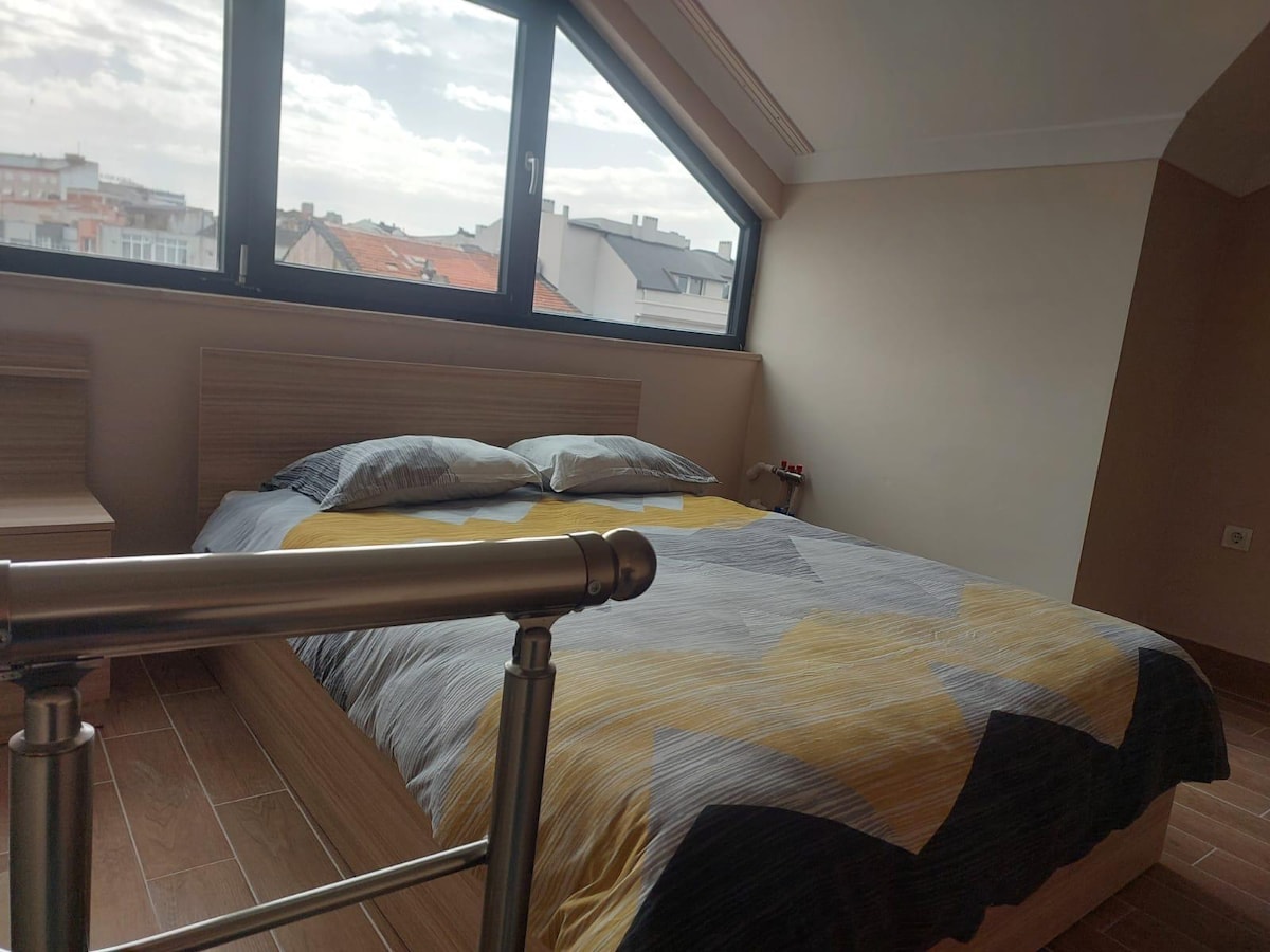 A bright sleeping area features a spacious bed with a geometric-patterned gray, yellow, and white blanket. Large windows provide ample natural light, complementing the minimalist wooden furniture on either side of the bed. The soft-colored walls create a calming environment.
