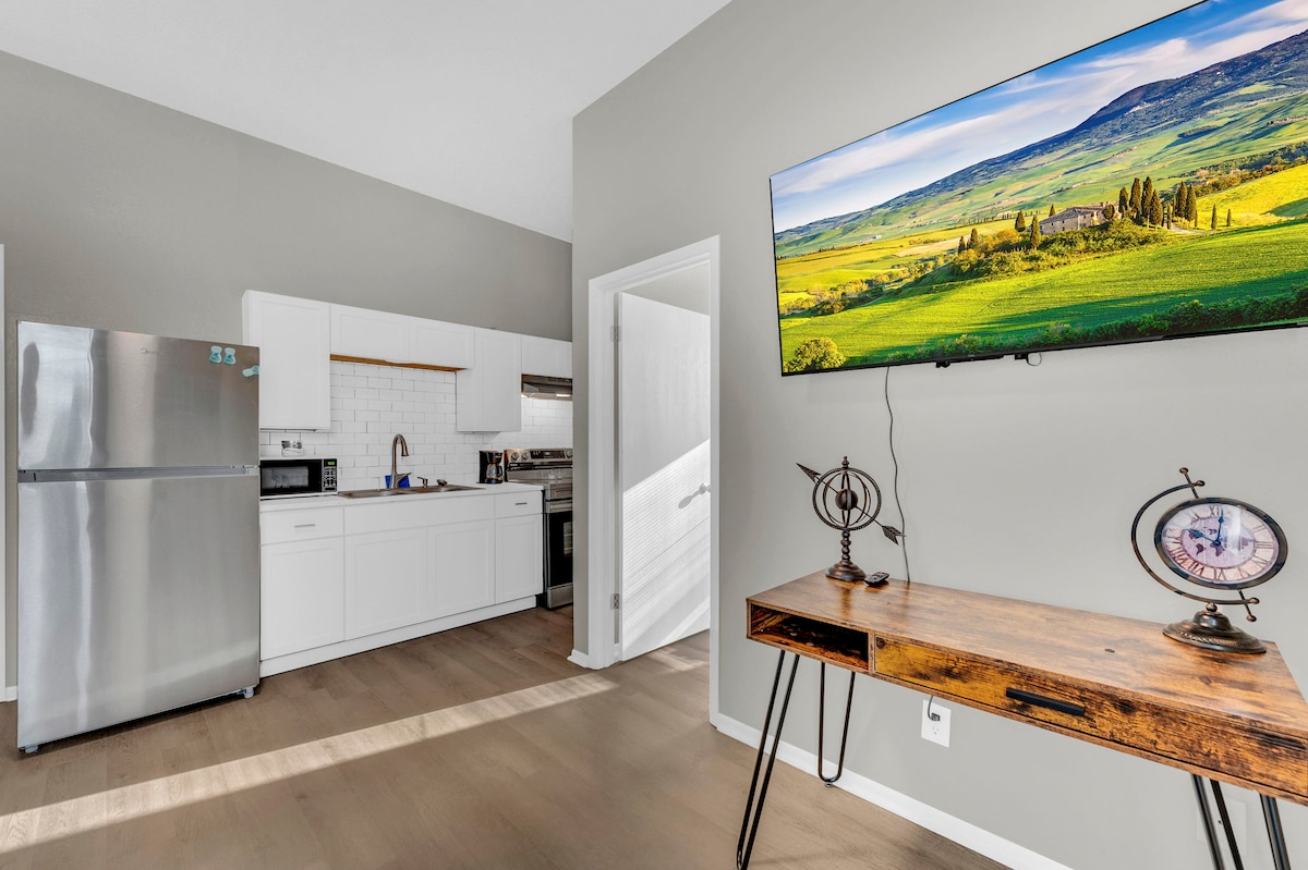 A bright kitchen area is visible, featuring modern appliances including a refrigerator and microwave. A wall-mounted flat-screen TV displays a scenic landscape. A rustic wooden table with decorative accents adds character, while natural light highlights the clean lines of the open layout.