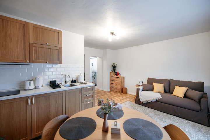 ღია გეგმარების მყუდრო მისაღები ოთახი თბილი ხის სამზარეულოთი, კომფორტული დივანი და სასადილო ზონა — იდეალური ადგილი დასასვენებლად ან ერთად სადილისთვის ✨