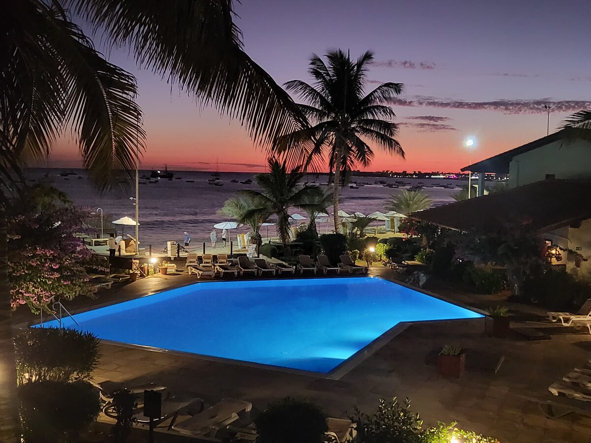 A tranquil swimming pool is illuminated in blue, surrounded by palm trees and lounge chairs. The sunset creates a colorful sky above the ocean, while a beach and umbrellas are visible in the background, enhancing the outdoor space's serene atmosphere.