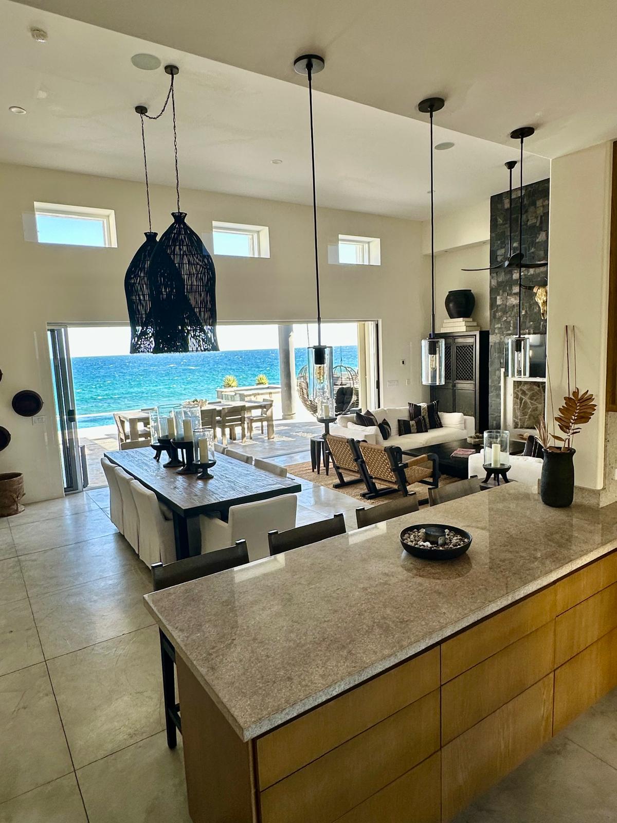 An open-concept living area showcases a modern kitchen with a central island, featuring natural stone countertops. The dining table seats several guests, while comfortable seating is arranged nearby. Large windows offer panoramic views of the ocean, allowing ample natural light to fill the space.